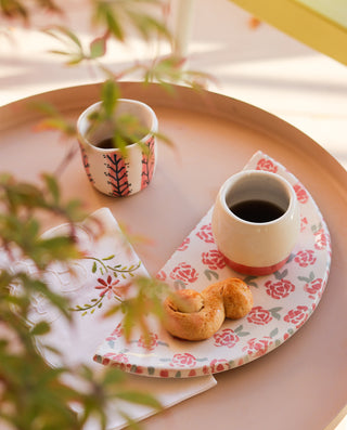 Two ceramic espresso cups with floral patterns, one with a pink base and the other with a white base, placed on a round tray with a matching design, accompanied by a white plate with a rose design, all set on a table with a blurred background.