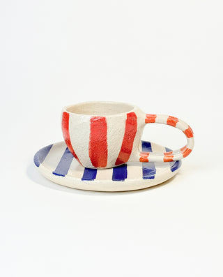 A hand-painted ceramic mug with vertical red and white stripes, accompanied by a round plate with horizontal blue and white stripes.