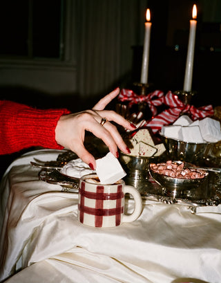 A hand with red nail polish places a white sugar cube into a red and white checkered mug on a draped white tablecloth. The setting is warm and festive with lit candles, a red striped ribbon, and bowls of chocolates or candies in the background.