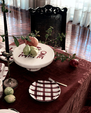 A styled hand painted dining table setting with a dark red tablecloth, a white pedestal plate holding green pears and a peach, patterned plates, a black candlestick, and a woven cane chair in a softly lit room.