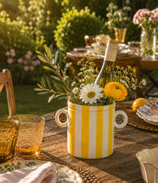 Decorative table setting with a yellow and white striped vase holding flowers and a bottle on a wooden table outdoors.