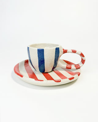A hand-painted ceramic mug with vertical blue and white stripes, accompanied by a plate with horizontal red and white stripes.
