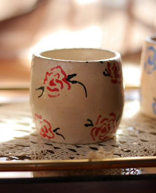 A beige ceramic stoneware mug with red floral pattern on it, placed on a lace tablecloth.