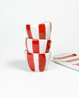 A set of three red and white striped espresso cups stacked together next to a matching plate, all placed on a white surface.