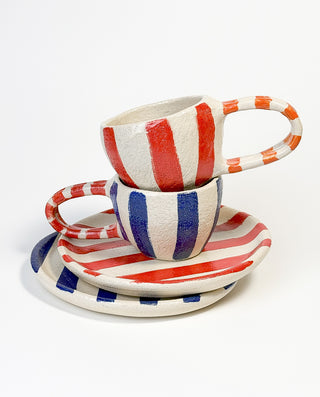 Two striped ceramic mugs with matching round plates, one set in blue and red stripes, the other in red and orange stripes.