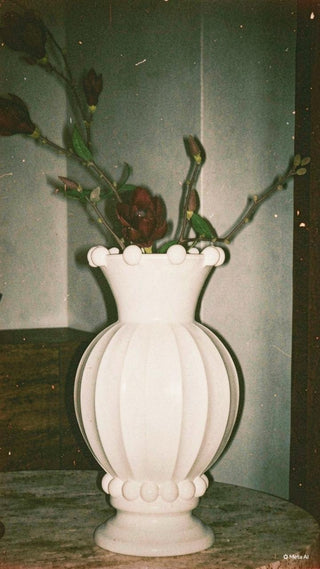 A white ribbed ceramic vase with a flared rim and decorative bead-like accents at the top and bottom, holding several dark rose stems, photographed indoors against a muted gray wall.