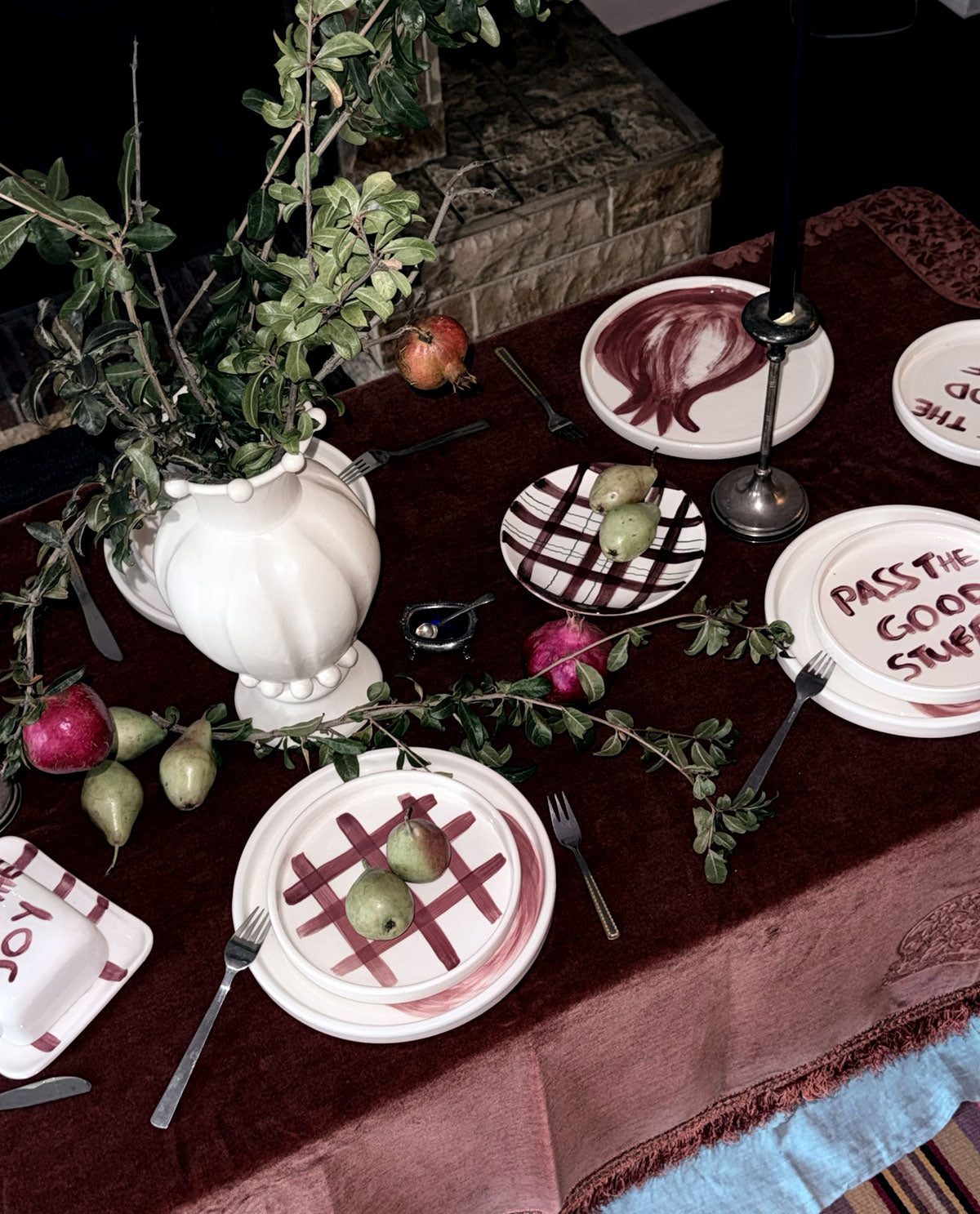 A styled dining table setting with white plates featuring red painted designs, silver cutlery, a white urn filled with green foliage, and pears scattered around on a dark red tablecloth.