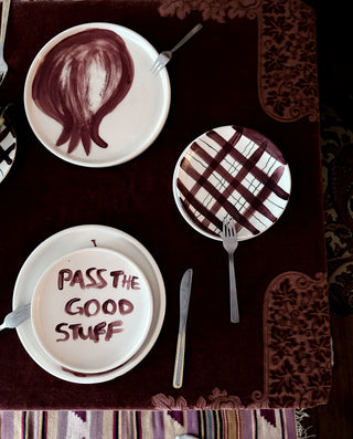 Three white ceramic plates with silver forks and a knife, arranged on a dark brown patterned tablecloth. The plates feature hand-painted designs: one with a dark brown hair-like swirl, one with a brown plaid pattern, and one with the text 'PASS THE GOOD STUFF' written in brown.