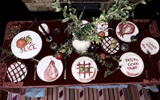 A styled table setting with multiple white plates featuring red and black food-themed illustrations and text, arranged on a dark red tablecloth with greenery and candles.
