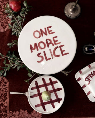A white round ceramic cake stand with the phrase 'ONE MORE SLICE' hand written , styled on a dark red velvet surface with a small plate, a green pear and decorative botanical elements.