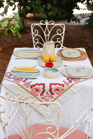 Outdoor table setting with decorative tablecloth, ceramic plates, and fruit on a white metal table and chairs.