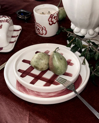 A styled tabletop scene with a white ceramic plate set featuring a dark red lattice pattern and painted red accents, holding two green pears and a metal fork. A matching candle holder and other tableware are visible on a deep burgundy tablecloth.