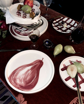 A tabletop dining scene with white plates featuring painted pear motifs, wine glasses, fruit, and a dark red tablecloth in a styled indoor setting.