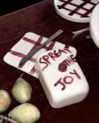 A white ceramic butter/cream container with 'SPREAD THE JOY' written in dark red lettering, displayed on a dark red velvet surface next to green pears and a matching checkered dish.