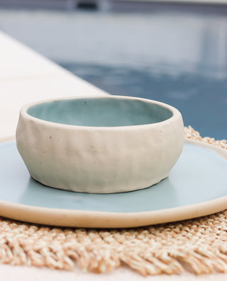 A handcrafted ceramic bowl with a sandy appearance, placed on a blue plate, with a pool and part of a white staircase visible in the background.