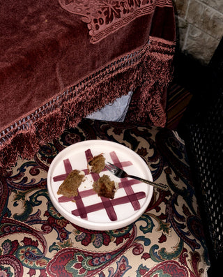 A white ceramic plate with a red grid pattern, containing two pieces of browned cake and a fork, placed on a patterned paisley chair cushion next to a dark red velvet draped fabric.