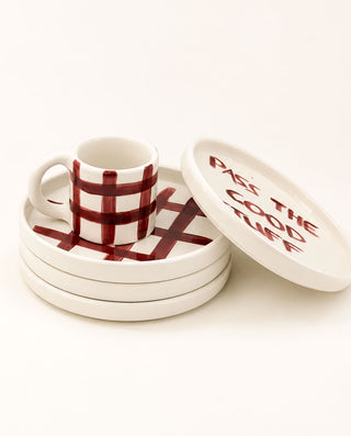 A white ceramic mug with a dark red plaid pattern sits on a matching stack of three coasters, with a round lid leaning against the top coaster. The lid features the text 'PASS THE GOOD STUFF' in red.