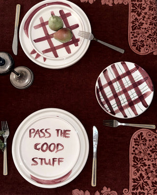 Three white ceramic plates with burgundy patterns, set on a dark red tablecloth with silver cutlery. One plate features the phrase 'PASS THE GOOD STUFF' in burgundy lettering, and two other plates display burgundy plaid and checkered designs.