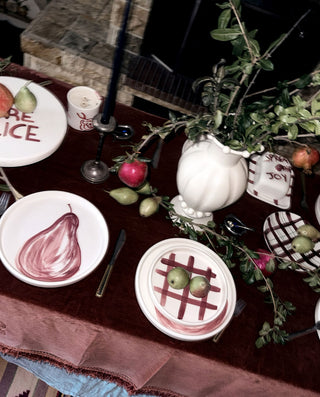 A styled tabletop setting with white hand painted ceramic plates by VECTORS & ROSES featuring painted pear and lattice designs, a white pitcher vase filled with greenery, red apples, olives, and a dark red tablecloth.