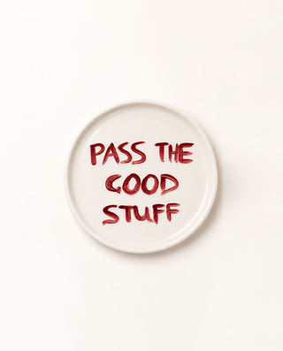 A round white ceramic plate with the phrase 'PASS THE GOOD STUFF' written in a textured red brush-style font, photographed from directly above on a clean white background.