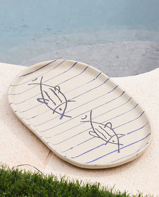 A beige oval serving platter with a hand-drawn fish and stripes design, placed on a grassy surface near a pool.