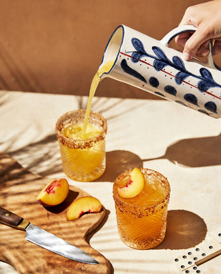 Person pouring a drink from a decorative ceramic pitcher into a glass with peach slices on a wooden surface.