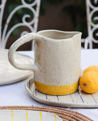 A beige and yellow ceramic handcrafted carafe with a colored glaze, placed on a table with a lemon beside it.