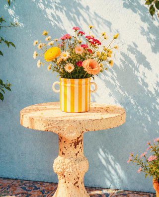 Yellow striped vase with flowers on a round stone table against a light blue wall.